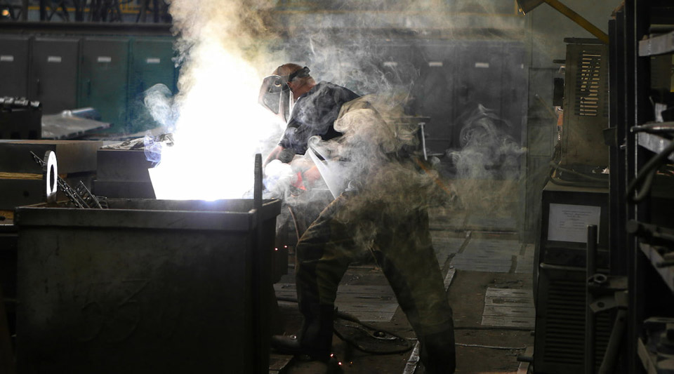 A welder wearing a protective mask and camouflage pants works on a metal container, surrounded by bright sparks and smoke.